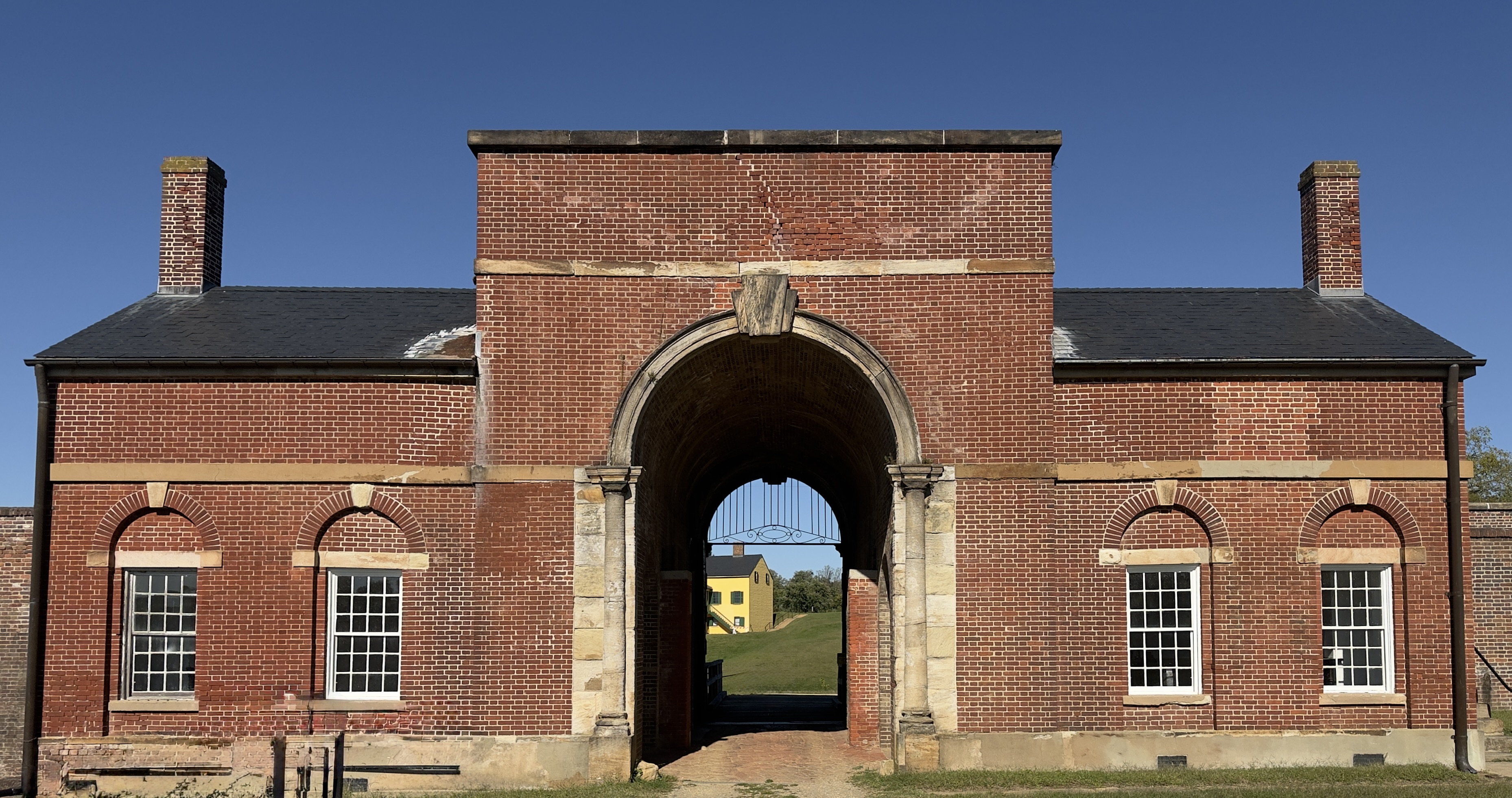 Symmetrical red-brick guardhouse at Fort Washington with a central stone arch, arched windows, and a glimpse of a yellow house through the gate
