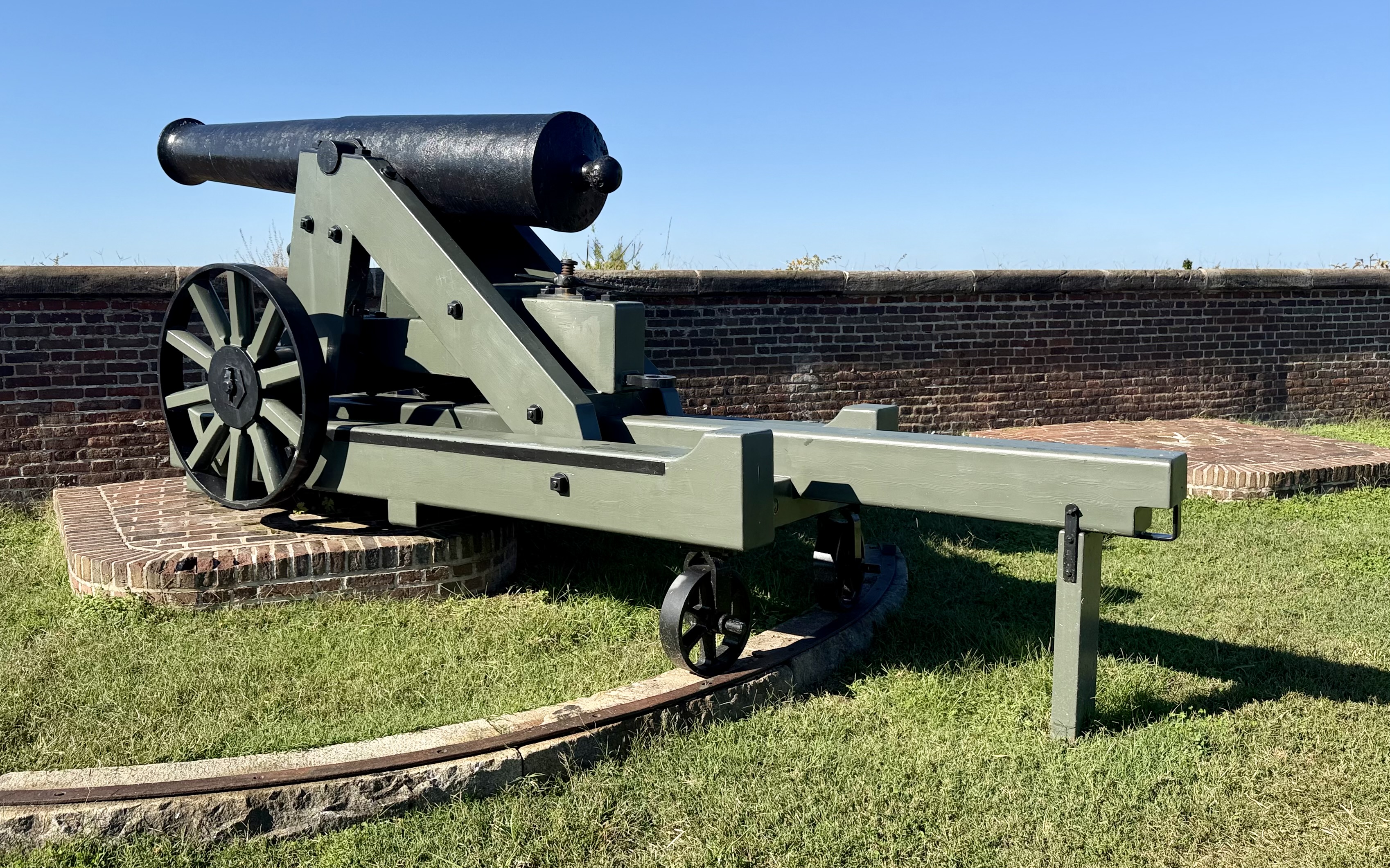 Green-painted Civil War–era cannon on a pivot track in front of a brick parapet wall under a clear blue sky