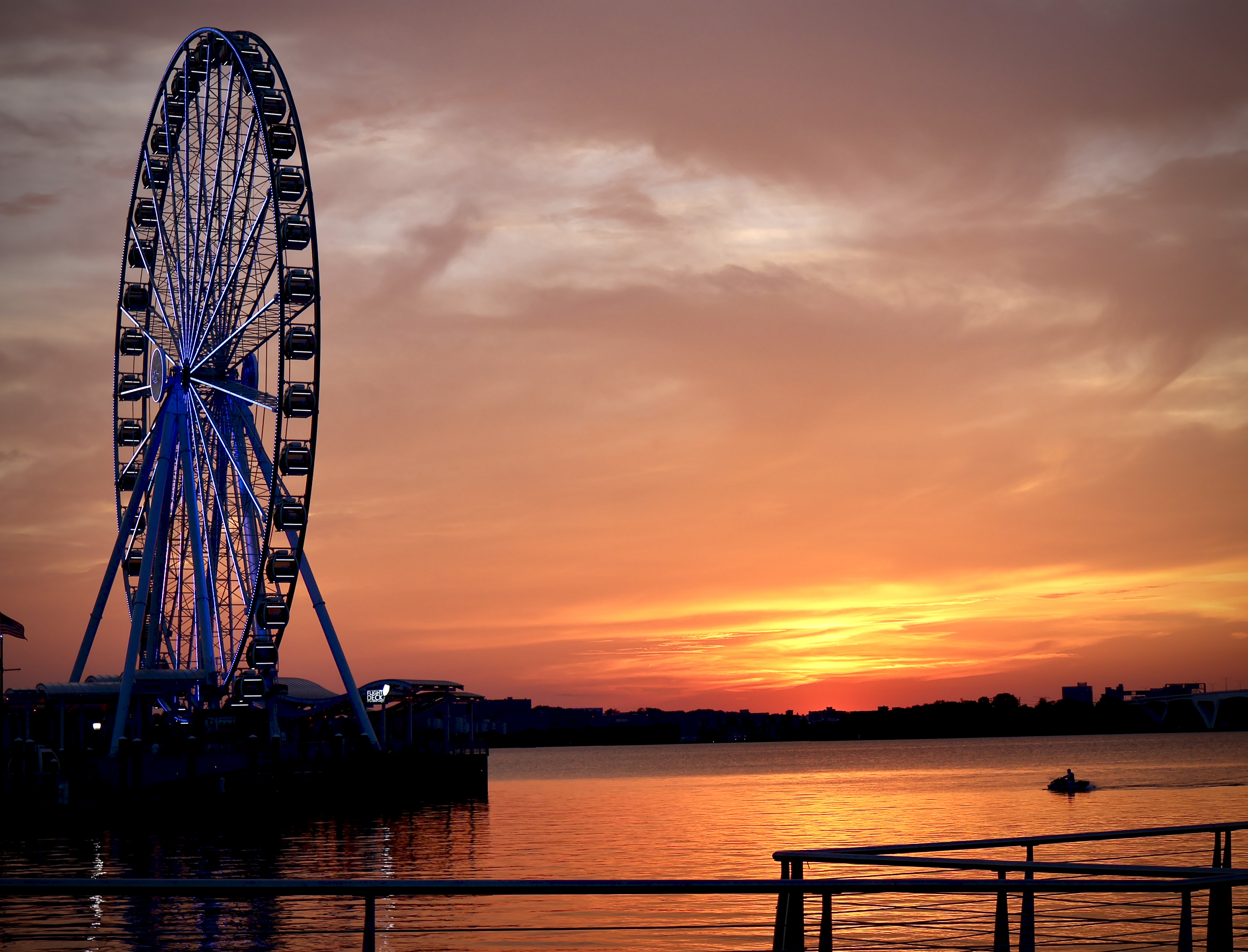 National Harbor Ferris wheel silhouetted against a deep orange sunset, seen past a pier railing with a jet ski passing on the water