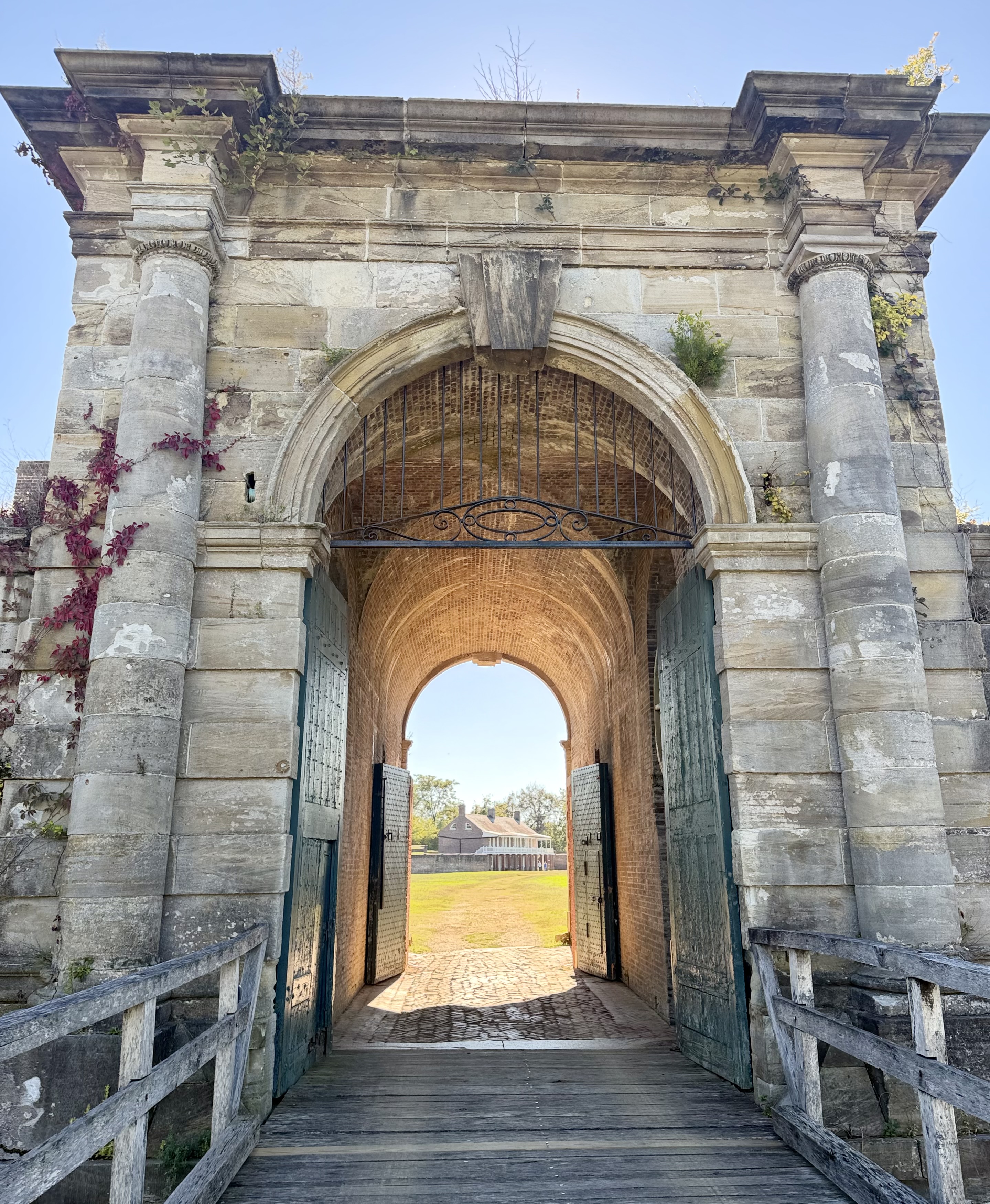 Stone gatehouse of Fort Washington with columns, ivy, and a wooden bridge leading through the arch to a distant house