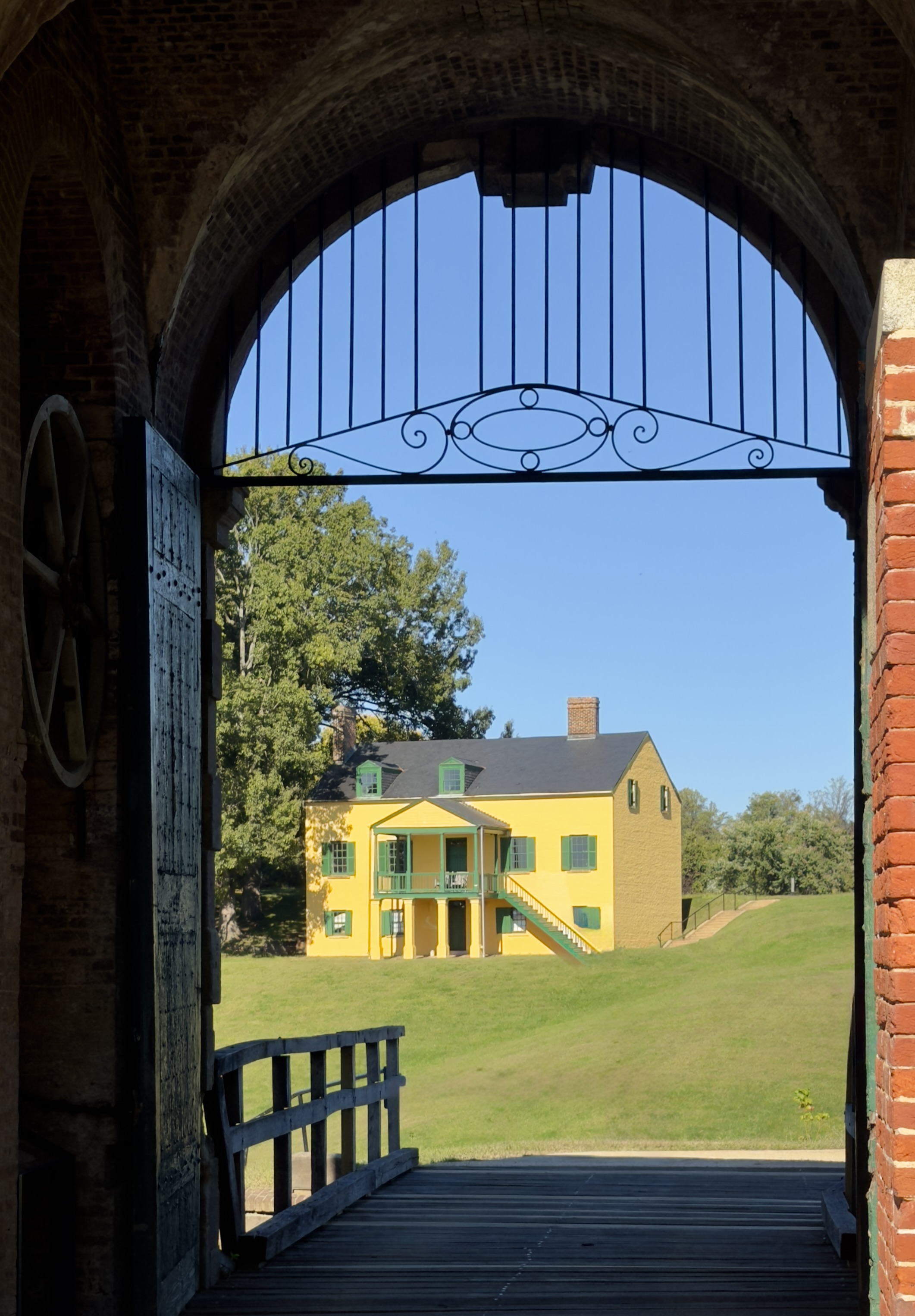 Yellow historic house seen through the iron-barred arch of Fort Washington's gate, with an open wooden door and a plank bridge in the foreground
