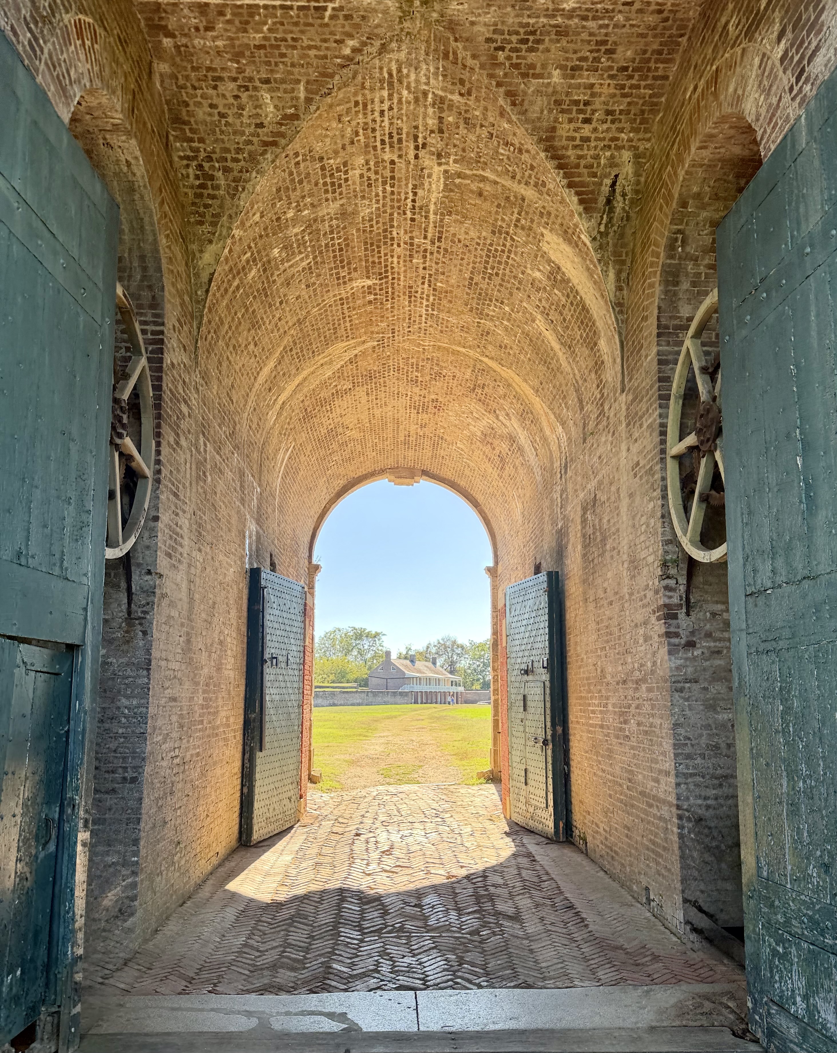 Looking down a sunlit brick-vaulted tunnel at Fort Washington toward a distant house, framed by open green doors and wooden gate wheels