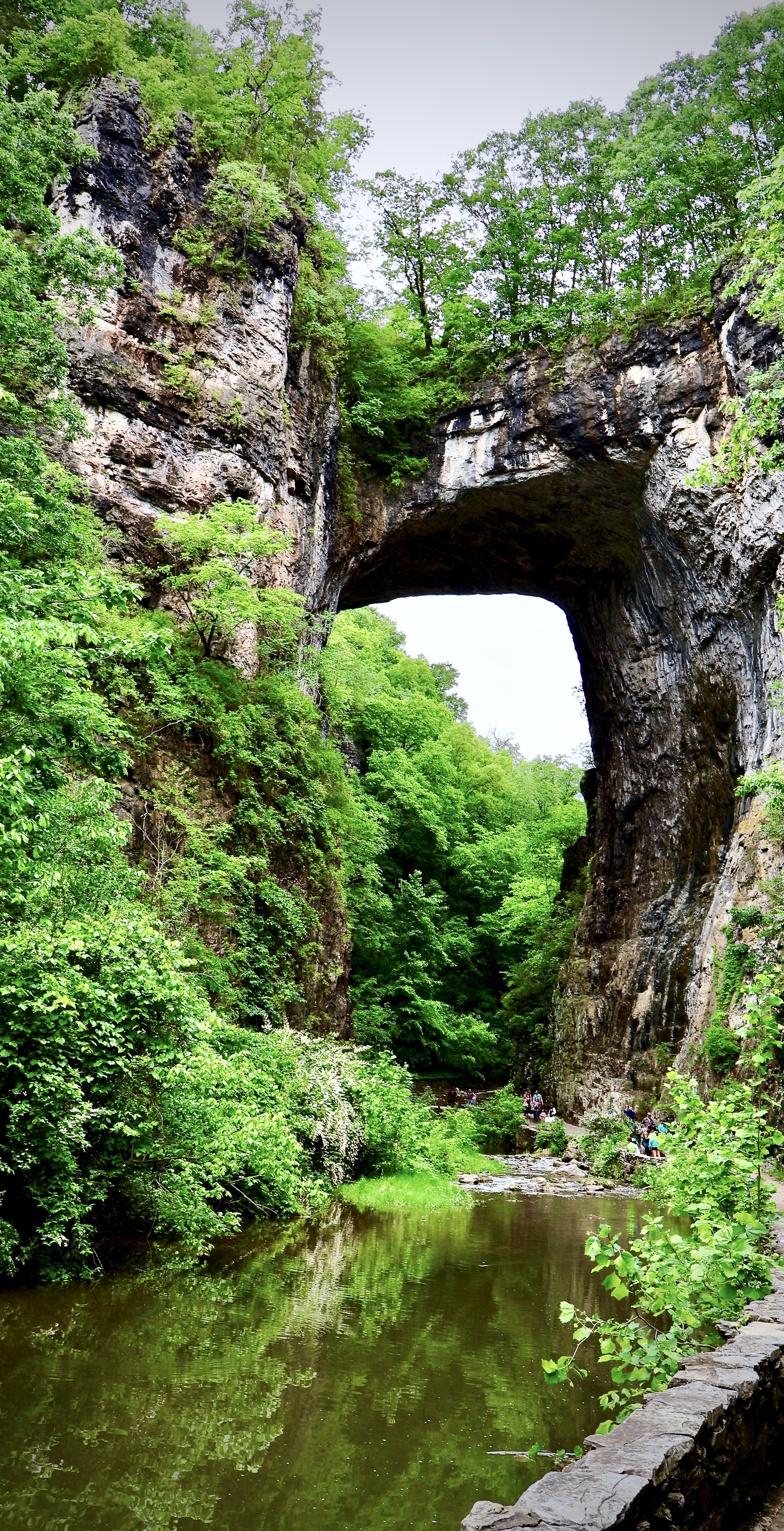 Natural stone bridge arch towering over a river, with its reflection in the water and people on the path below