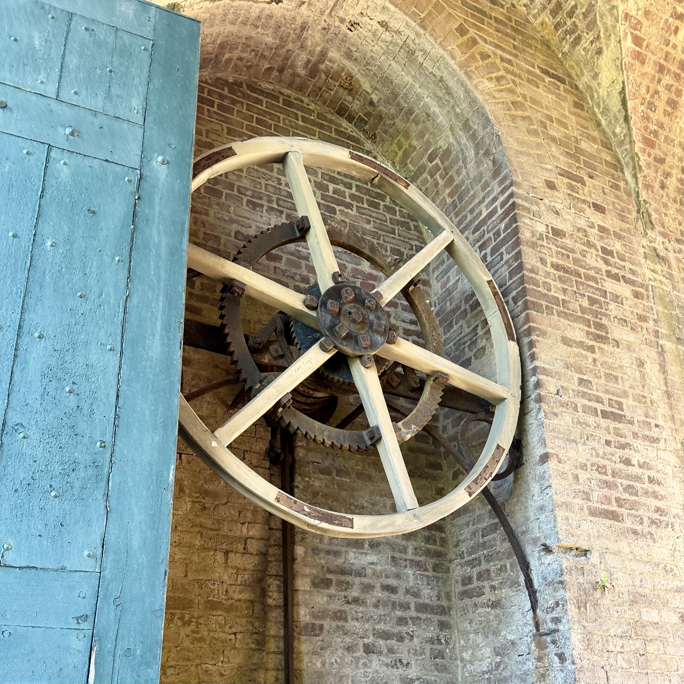 Large wooden gate-winding wheel and iron gears mounted on a brick wall beside a weathered blue door